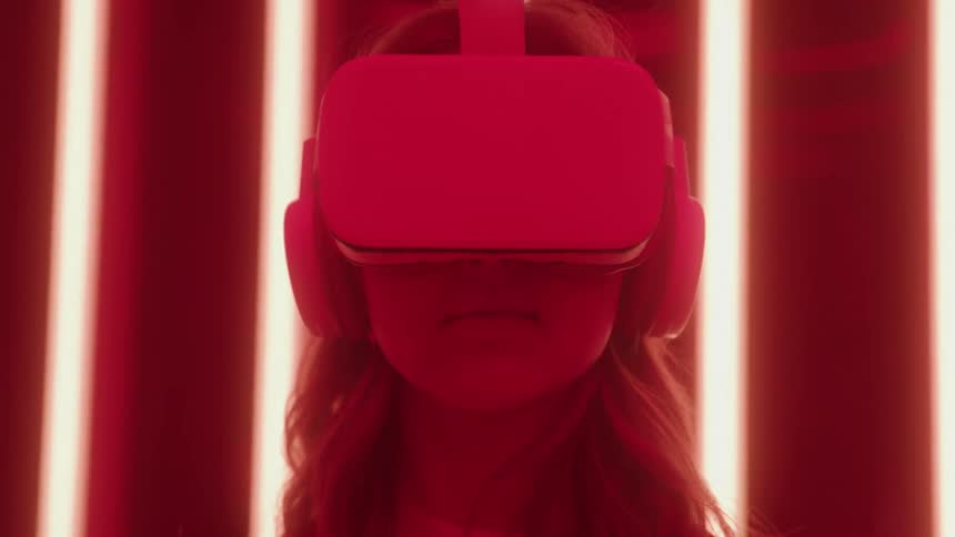A woman with long wavy hair wears a virtual reality headset and headphones while illuminated by bright vertical red lights in a dark room