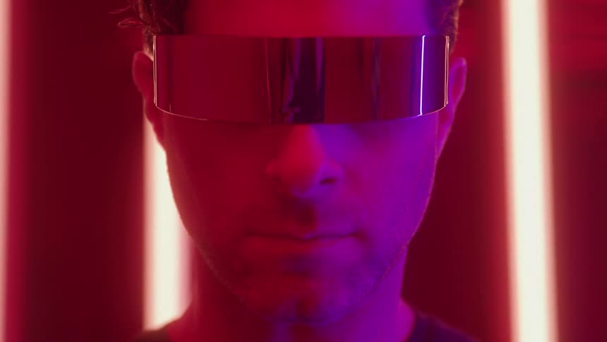 A closeup of a man with a slight smile looking forward while wearing futuristic mirrored visor glasses illuminated by strong red and purple neon lights in a dark background