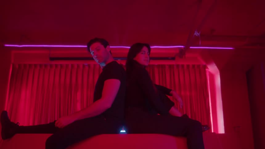 A man and a woman in black clothes sit back to back on a red surface in a club illuminated by intense pink and red neon light