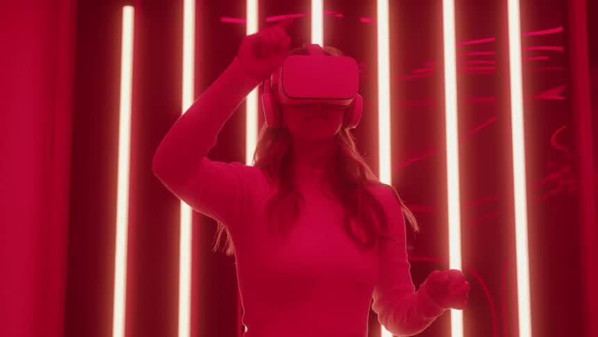 A young woman wears vr glasses in a room illuminated by red led lights using her hand gestures to control the device