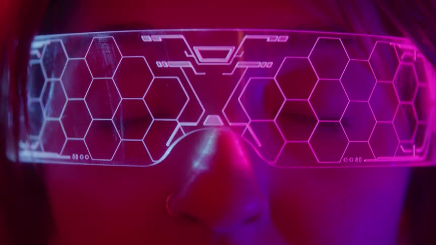 A closeup of a persons eyes and nose with a neutral expression looking forward while wearing clear futuristic glasses illuminated by vibrant pink and purple neon light