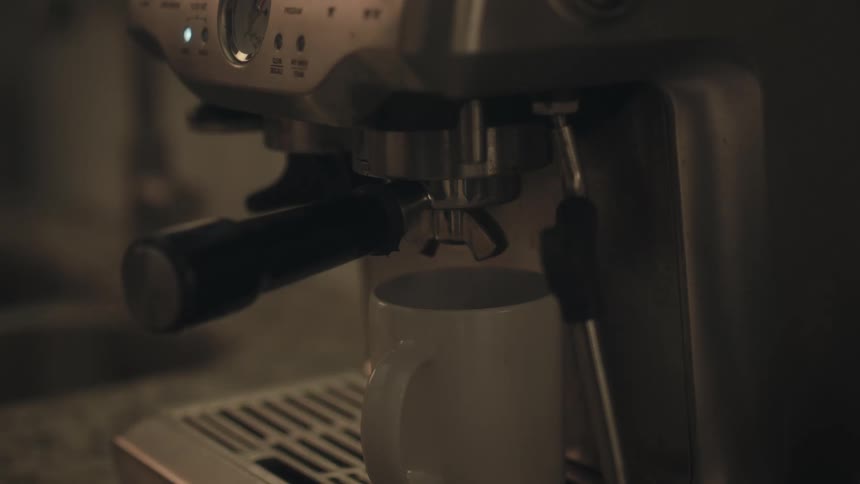 A stainless steel espresso machine shows the dark liquid coffee being extracted from the portafilter and slowly dripping into a white ceramic mug placed on the machines drip tray in a dimly lit setting