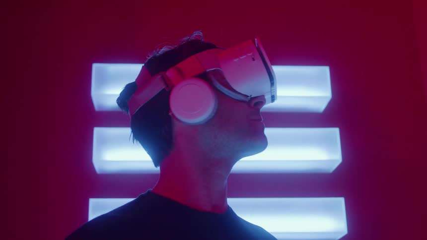 A man wearing a white virtual reality headset and headphones looks around an indoor space illuminated by red and bright horizontal blue neon lights