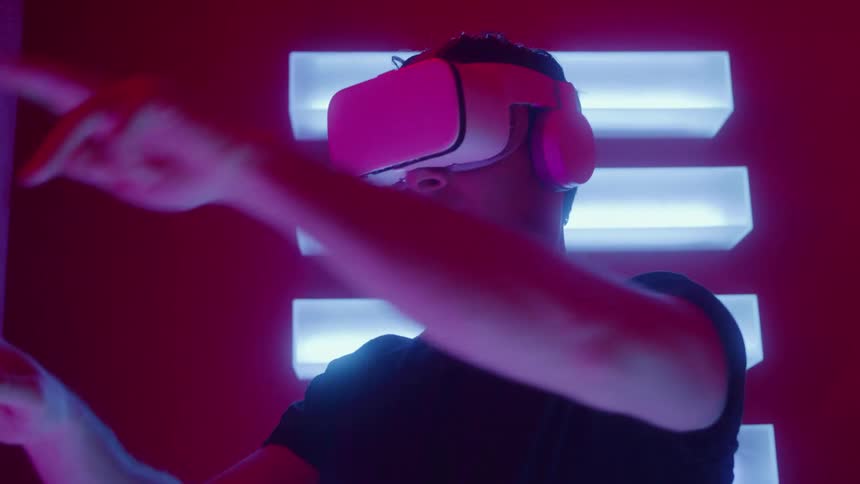 A man in a black shirt wears a white virtual reality headset and headphones while interacting with an unseen interface in a dark room illuminated by neon pink and blue light