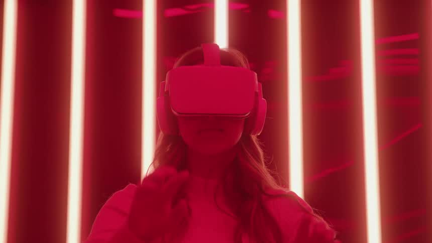 A medium close view of a woman wearing a white sweater and a virtual reality headset reaching out her hand in front of glowing vertical neon red lights