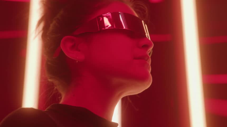 A woman wearing dark mirrored visor glasses is shown in a tight lowangle profile closeup illuminated intensely by a deep red light from vertical neon tubes