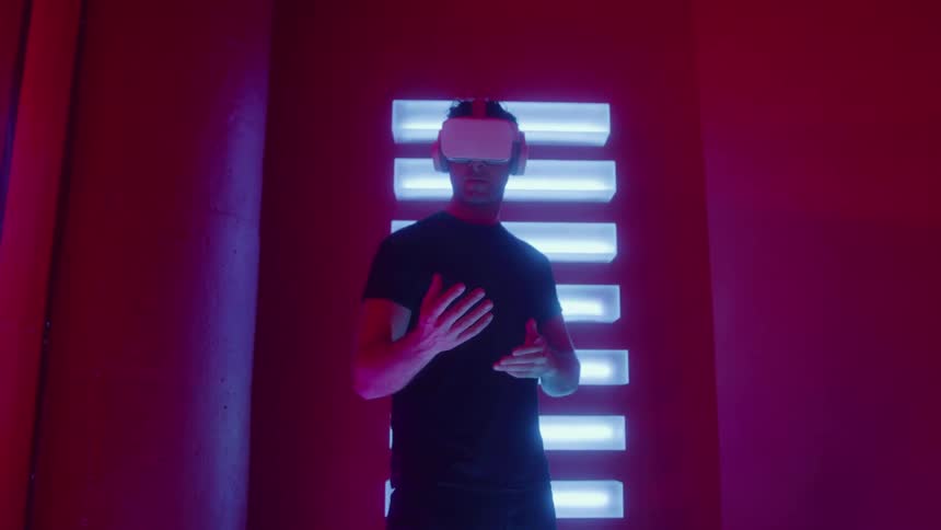 Young adult man wearing a virtual reality headset and looking around in a colorful room with neon lights indoors