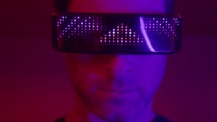 A closeup shot of a mans face in dark neon red and purple lighting shows him wearing a pair of futuristic glasses with an active led display which cycles through a bright colorful pixelated pattern