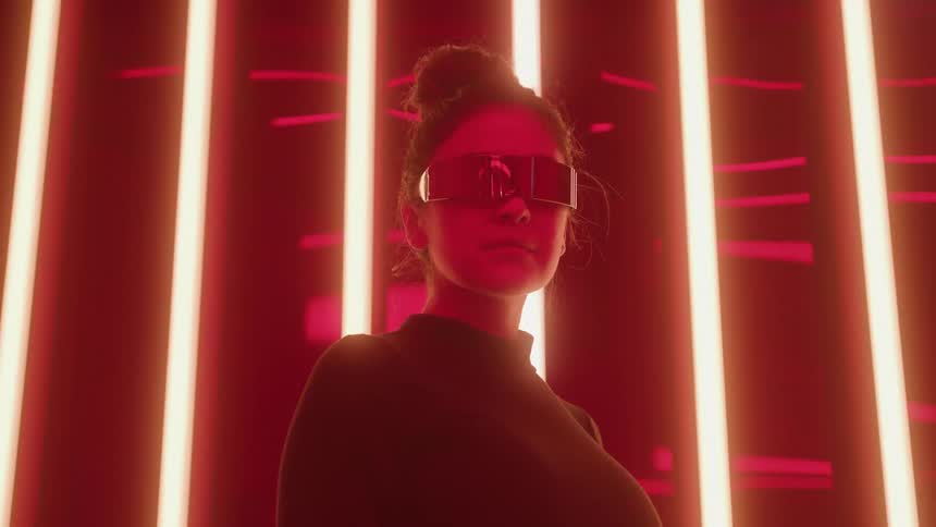 A woman wearing futuristic mirrored rectangular glasses and a dark top is illuminated by vertical neon red lights in the backgroundshe slowly turns her head to face the camera creating a cyberpunk or scifi aesthetic
