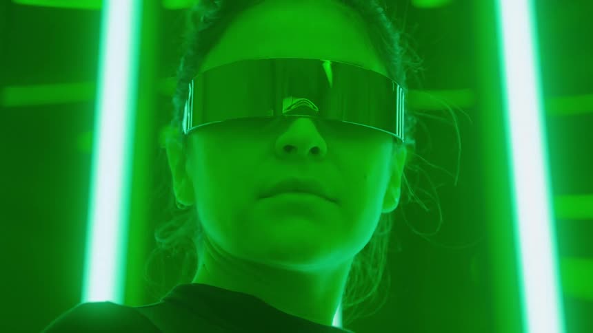 A woman wearing large mirrored visor glasses and a dark top is shown in a tight lowangle closeup illuminated by an intense vibrant green light emanating from vertical neon tubes