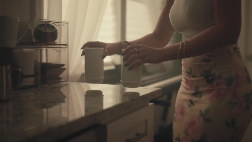 A woman wearing a white top and a floralpatterned garment as she walks toward a kitchen counter and places two identical white cups onto the glossy surface near clear storage containers and a window