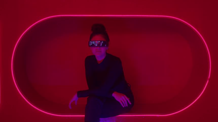 A mediumwide shot shows a woman dressed entirely in black wearing digital glasses with an active display sitting passively within an oval boothlike structure outlined and intensely lit by neon pink light