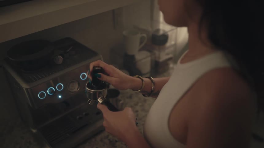 This is an overtheshoulder medium shot of a woman in a white tank top preparing coffee using a stainless steel espresso machine in a dimly lit kitchenshe is seen tamping down the grounds in the portafilter handle with the machines blue indicator l