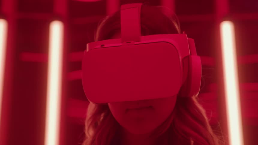 A young woman wears a virtual reality headset and integrated headphones engrossed in the experience under intense red lighting from vertical neon tubes in the background