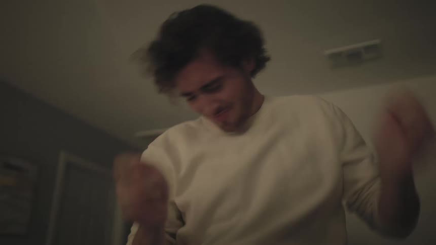 A young man in a white sweatshirt and jeans is sitting on a countertop happily dancing and moving his arms indoors in a dimly lit space