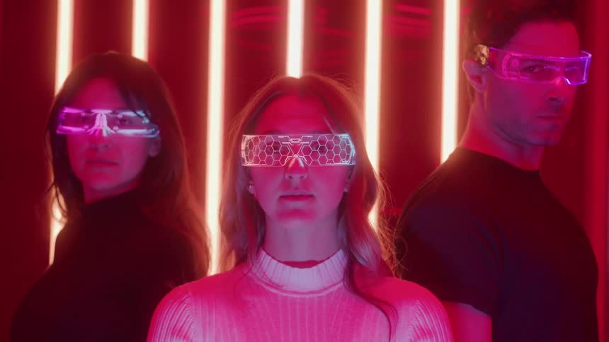 Three people wearing futuristic glowing visors and dark clothes stand together under bright red neon lights in a medium close view