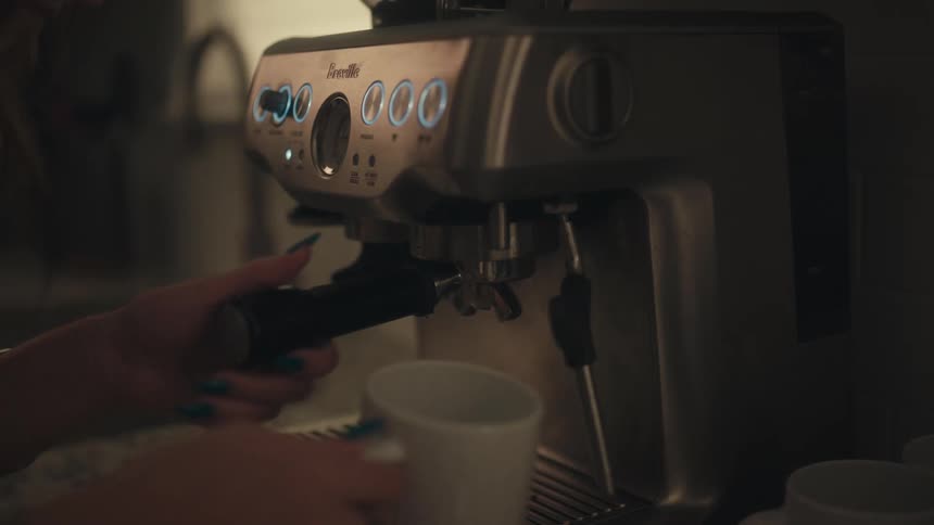 A close view of a persons hands with blue painted nails operating the portafilter and placing a white mug in a silver espresso machine in a dimly lit kitchen