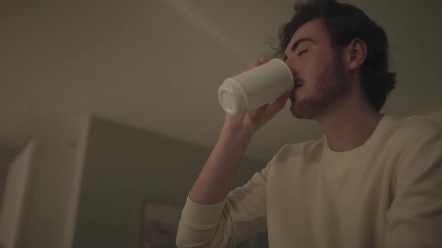 A young man with dark wavy hair and light facial hair is seated casually on a kitchen countertop in an indoor space smiling as he lifts a white mug to his lips to take a drink before lowering it and looking offcamera with an expression of satisfaction