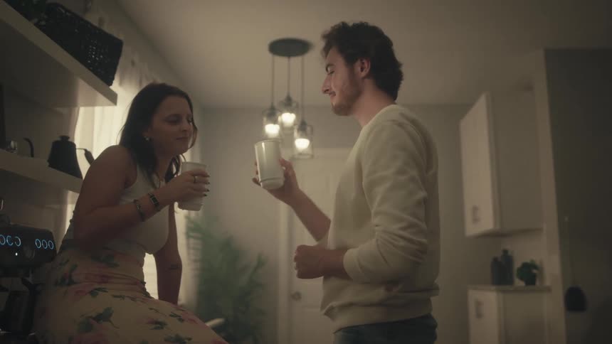 The video starts with a young man and woman in a dimly lit indoor space possibly a kitchen or studio apartment where the man is seated and taking a drink from a white mugthey then both stand up move closer clink their mugs together in a toast and