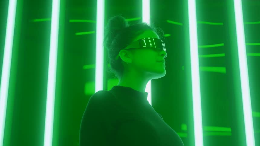 A woman wearing mirrored futuristic glasses and a black top stands in profile against vertical glowing green neon lights in a dark room