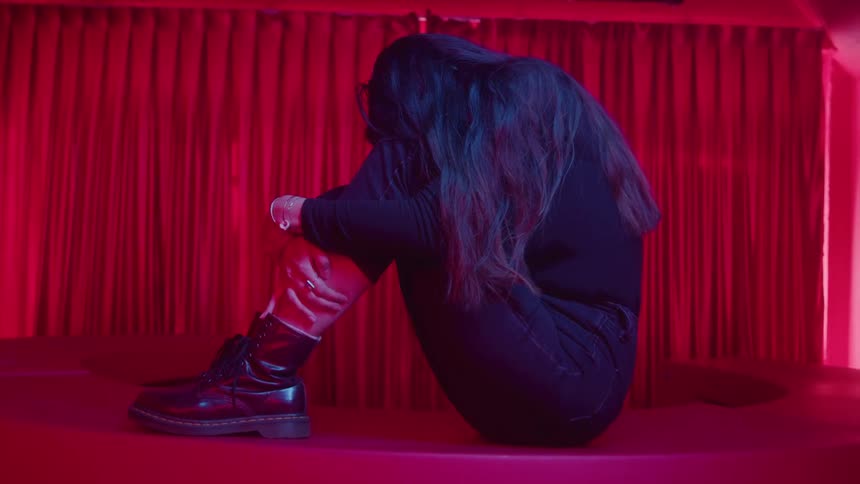 A woman with long dark hair wearing black clothing and laceup boots is sitting huddled on a red platform or stage seemingly distressed the scene is dominated by a strong red lighting with red velvetlike curtains serving as the background