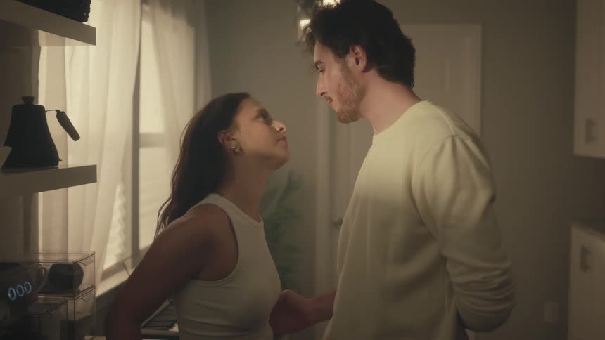 A young man and woman are standing close together in a softly lit kitchen leaning in toward each other and maintaining intense eye contact while engaged in a conversation and the man kisses the woman forehead