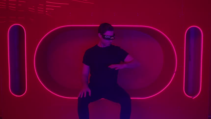 A man wearing dark clothes and futuristic glasses sits looking at his arms with a serious expression looking forward against a dark background illuminated by horizontal and vertical pink neon oval shapes