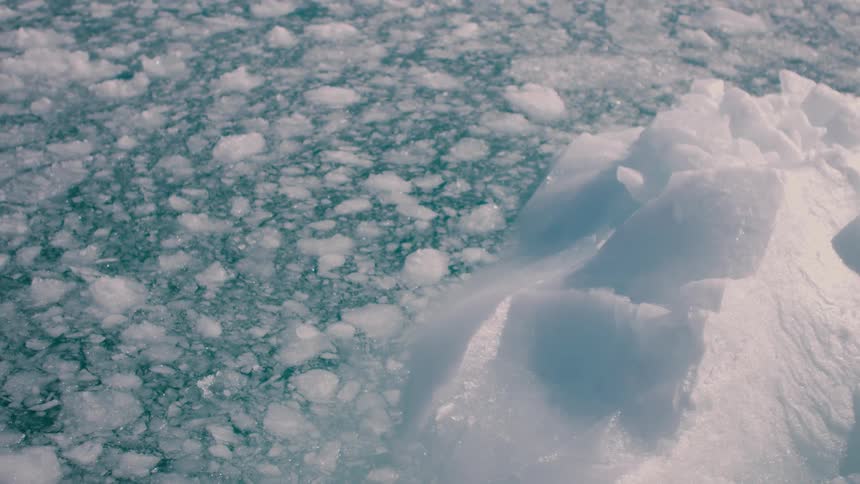 A close view shows the bright white textured surface of an iceberg piece floating near a large expanse of broken ice and slush on a cold blue ocean