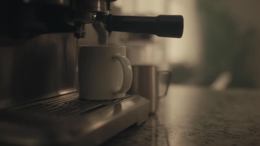 A persons hand places a white mug on a tray beneath the portafilter of a silver espresso machine as dark liquid drips into the cup in a dimly lit kitchen