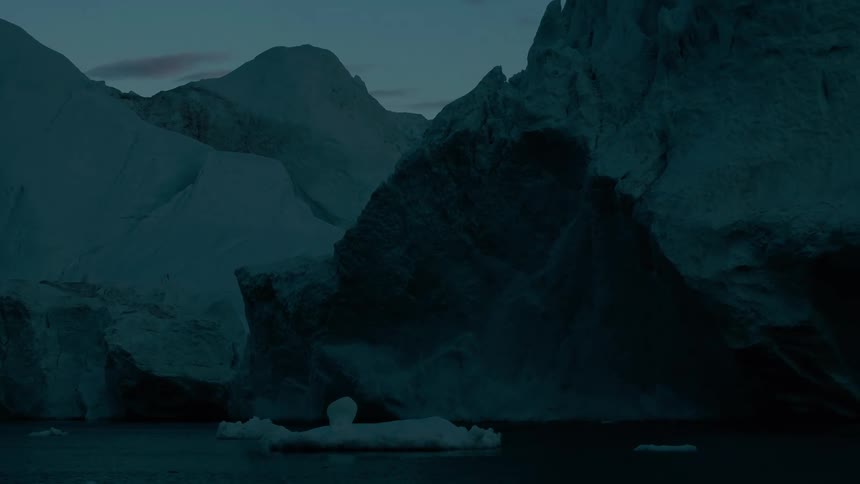 Towering jagged ice cliffs and massive glacial formations loom over dark deep water in a polar environment with scattered chunks of small icebergs floating in the foreground under a twilight sky