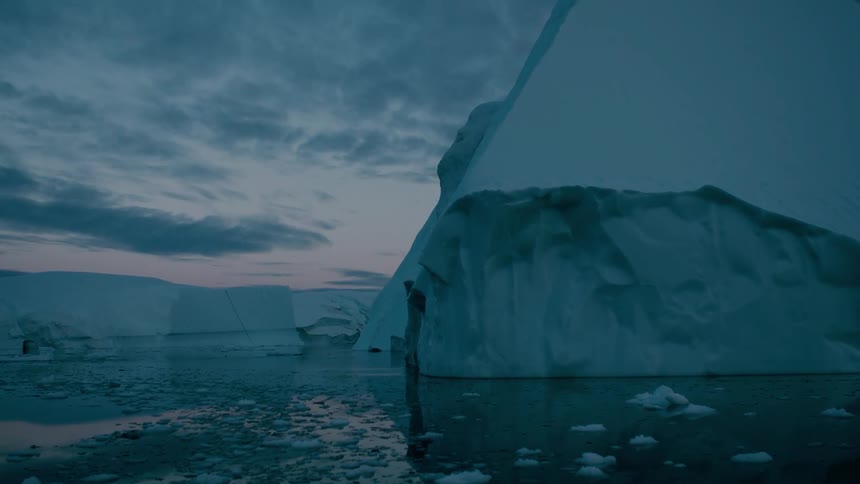 A massive angular iceberg floats on dark water surrounded by smaller ice chunks under a dramatic gray and blue cloudy sky at twilight
