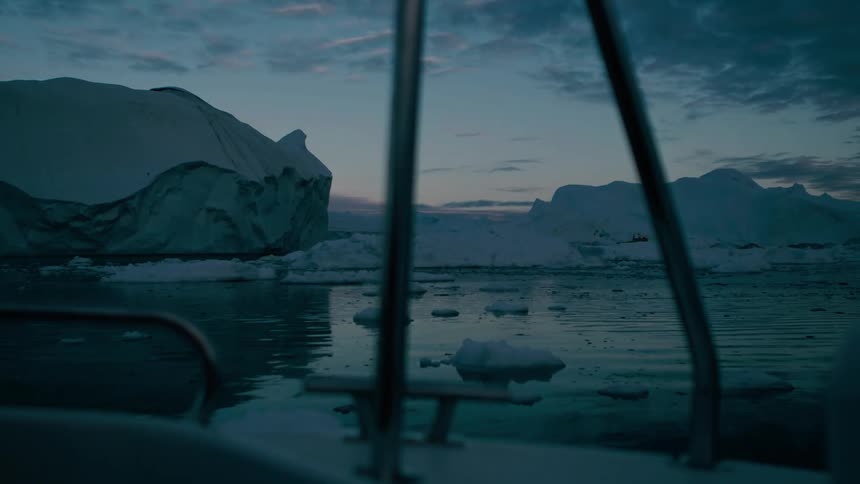 A view from a boat shows massive icebergs and floating ice chunks on dark water under a deep blue and pink twilight sky