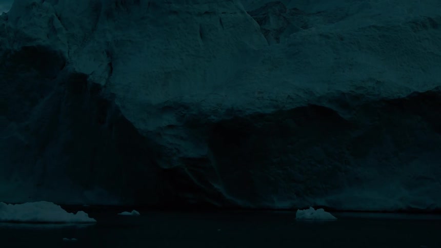This is a dark wide shot of a massive foreboding glacier or ice cliff towering over still dark water under low lightsmall icebergs float in the water below the dramatic textured face of the ice formation emphasizing its immense scale and the chill