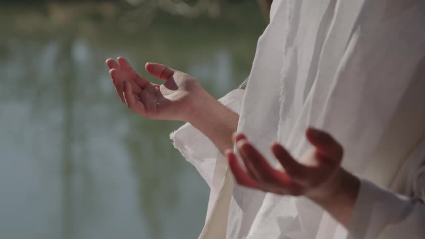 A woman in a white gown stands outdoors with her hands spread apart praying