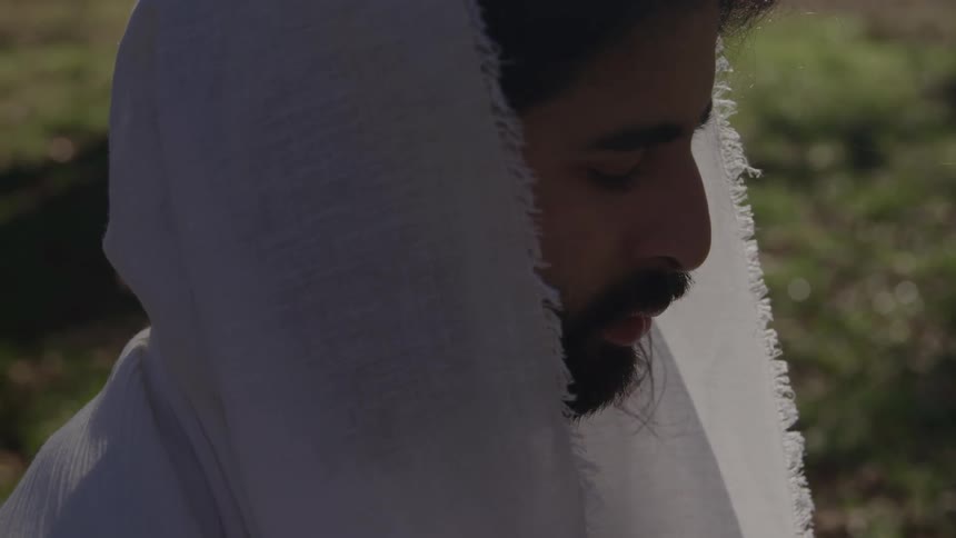 A man with a dark beard and mustache wearing a white hooded or draped cloth over his head is shown in a tight side profile closeup gazing downward in an attitude of contemplation in an outdoor sunlit setting