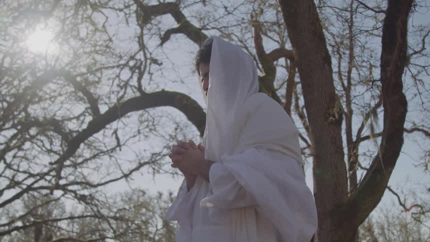 Man wearing a light hooded robe holding his hands together in a wooded area