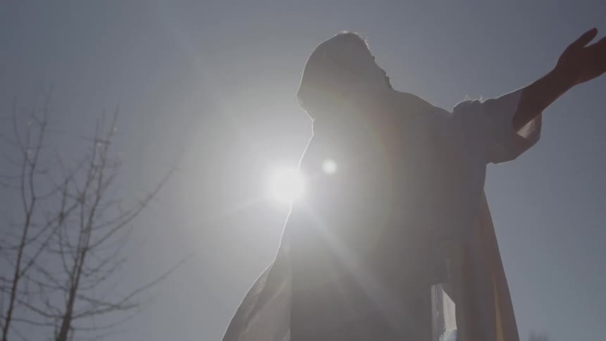 A backlit silhouette of a hooded figure in a white robe raising both arms toward the bright sun in a dramatic outdoor setting
