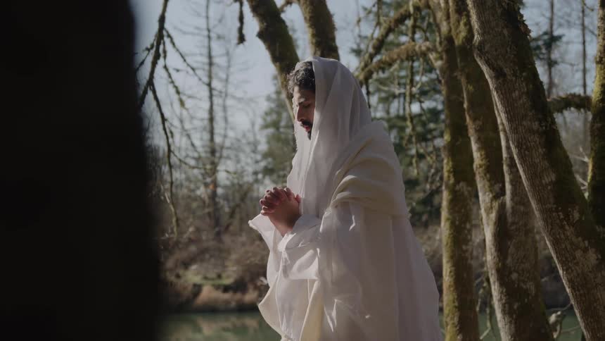 A man with a beard wearing a simple white robe and cowl stands in a mossy forest beside a body of water with his hands clasped in prayer