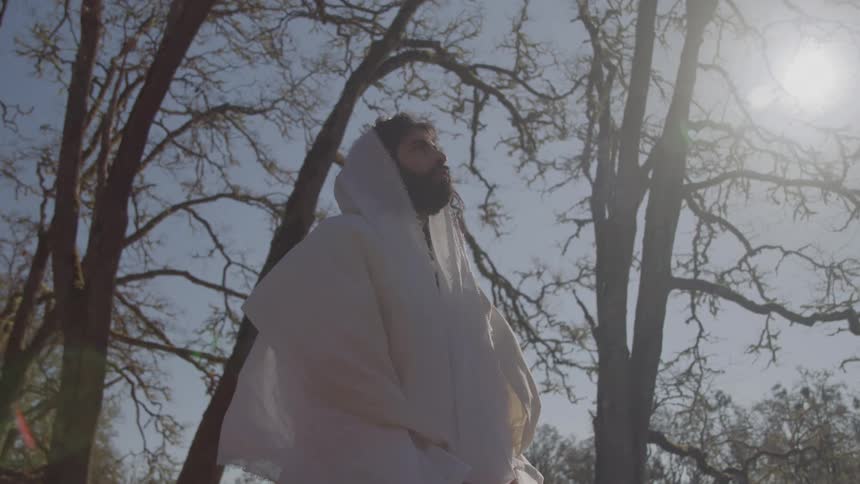 Man in a white hooded robe prays outdoors under bright sunlight