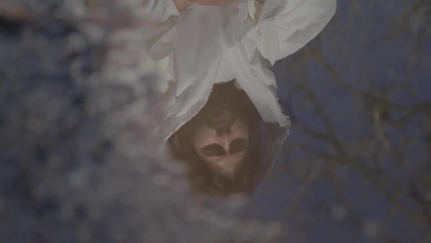 A close view shows the inverted and blurry reflection of a man in a white robe looking into a puddle with the surrounding trees reflected in the surface