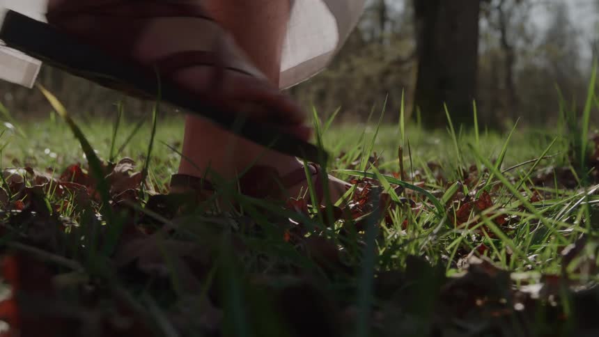 A closeup of a persons feet wearing brown leather sandals stepping on a patch of grass and fallen leaves showing an outdoor environment
