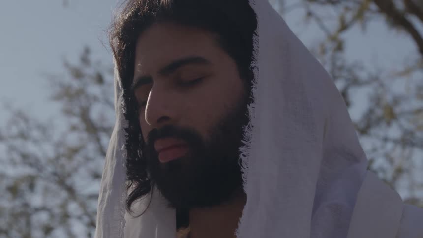 A man with a dark beard and long hair wearing a white hood or head covering has a meditative expression with his eyes closed looking forward outdoors