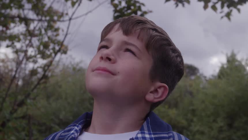 A teenage boy stands in the woods at midday smiling as he gazes up at the sky