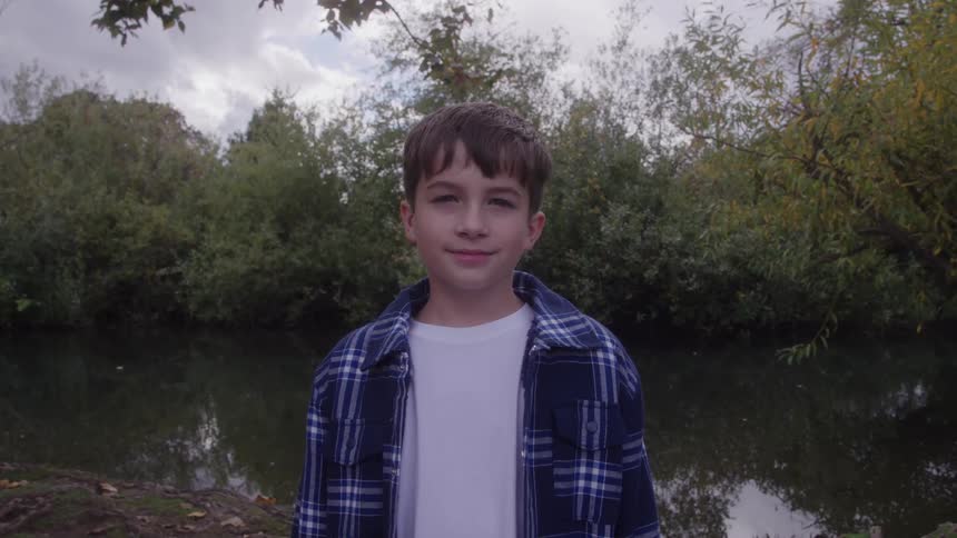 Young boy wearing a plaid blue shirt standing next to a pond next to a forest area outdoors during day time