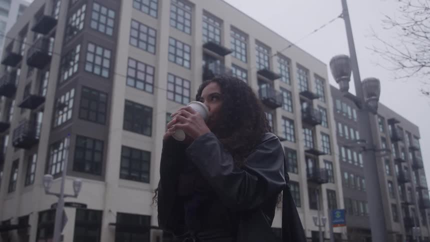 A woman enjoys coffee outdoors on an overcast day standing in front of a modern city building