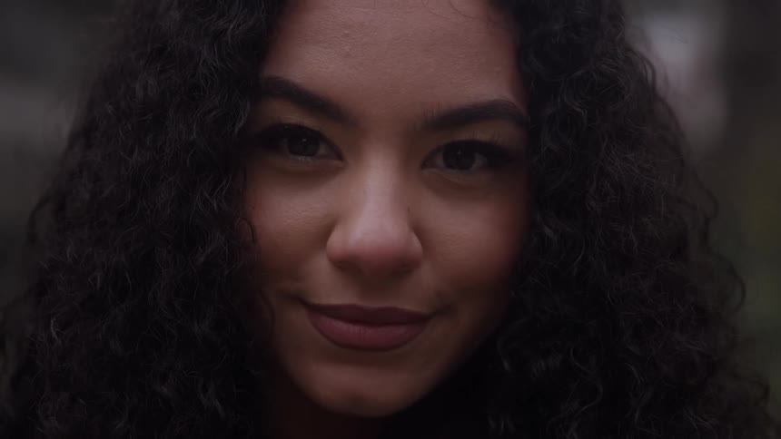 A woman with dark curly hair looks off to the side she turns her gaze forward with a warm gentle smile