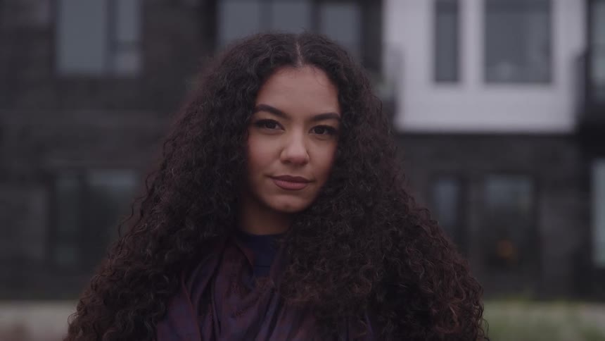 A young woman with long voluminous dark curly hair as she turns her head from a side profile to face the camera giving a direct and subtle smile against the blurred background of a modern dark building