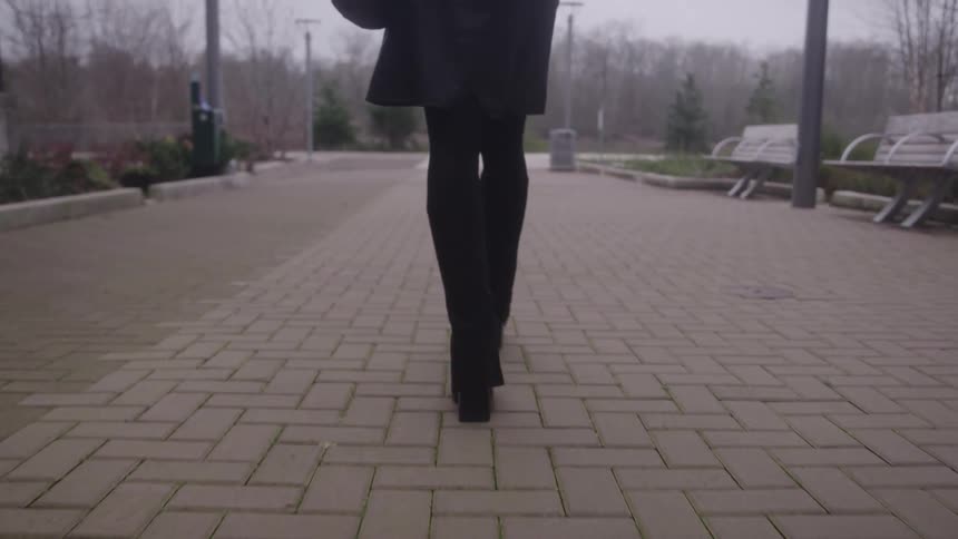 A low angle rear view shows a woman wearing black leggings and heeled boots walking away along a brick paved path in an urban park area