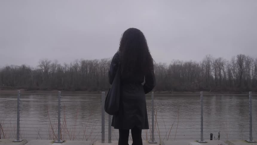 A woman with long curly hair stands with her back to the camera looking out over a wide river or body of water toward a wooded bank on a gray overcast day