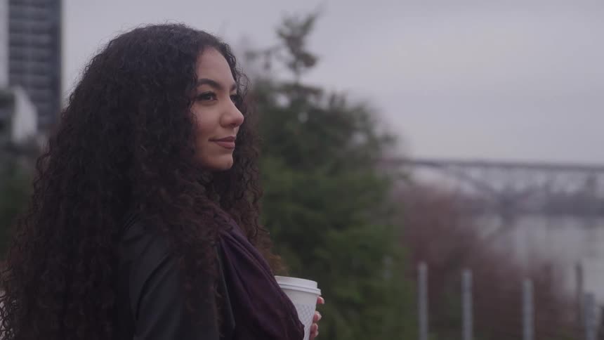 A young woman with very long dark curly hair stands outdoors on an overcast day looking off into the distance she is holding a white paper coffee cup and wearing a dark jacket with a purple scarf